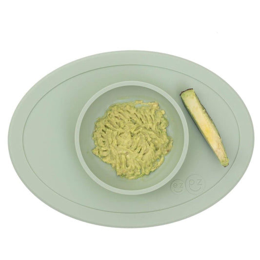 A sage green silicone toddler bowl with a raised central dish holding mashed avocado, with a cucumber spear placed alongside, viewed from above.