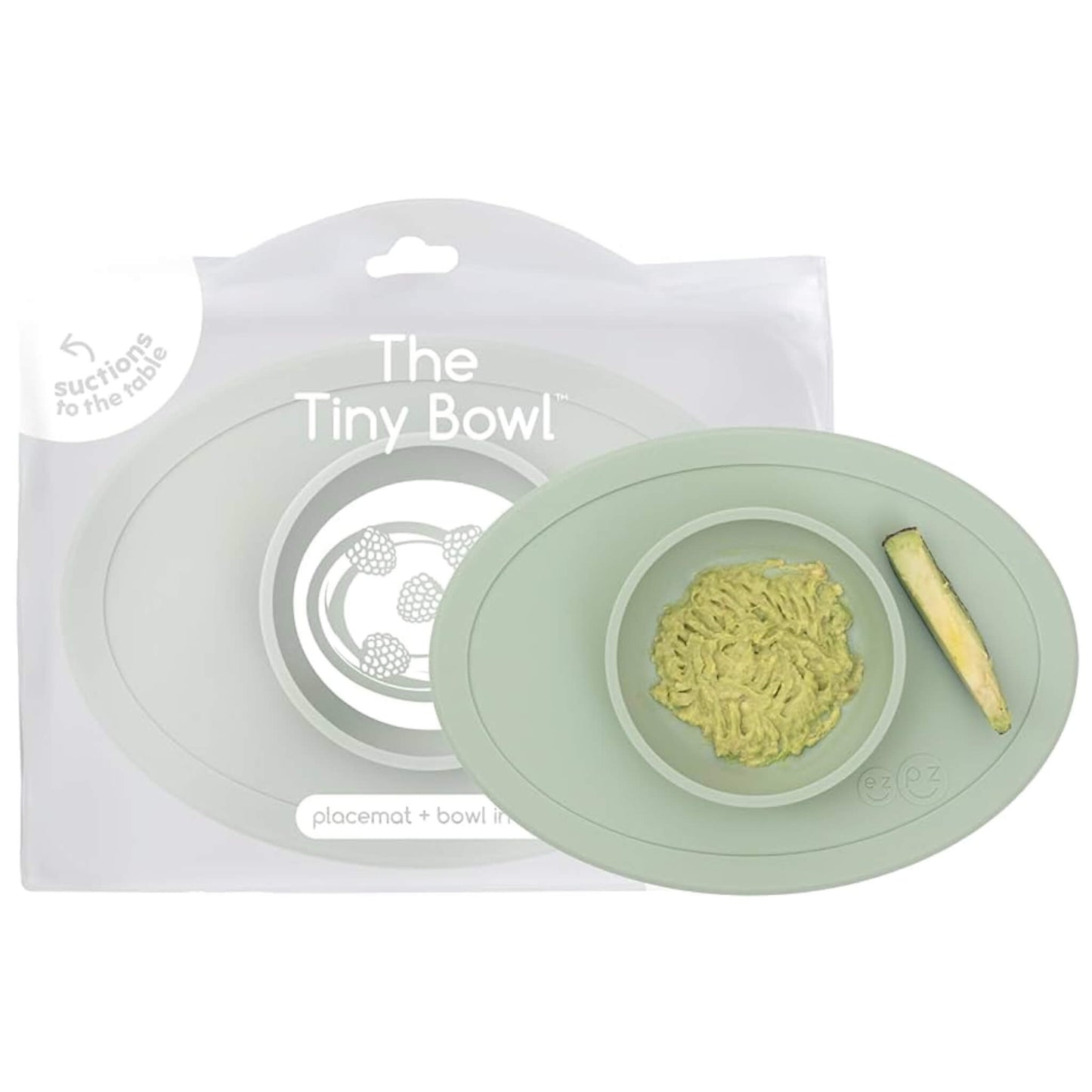 A sage green silicone toddler bowl shown with retail packaging behind it, featuring mashed avocado in the central dish and a cucumber spear to the side.