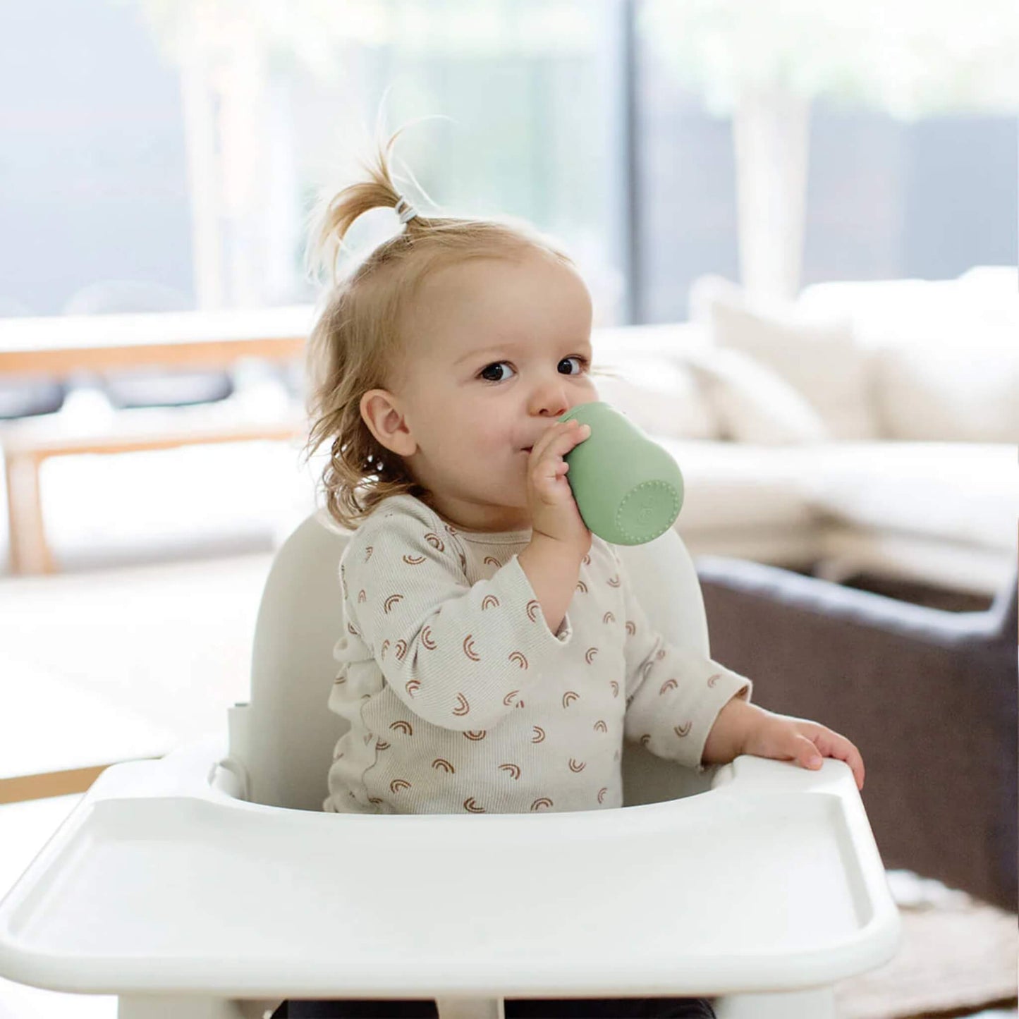 Toddler drinking from a sage green silicone straw cup while seated in highchair, demonstrating independent drinking skills