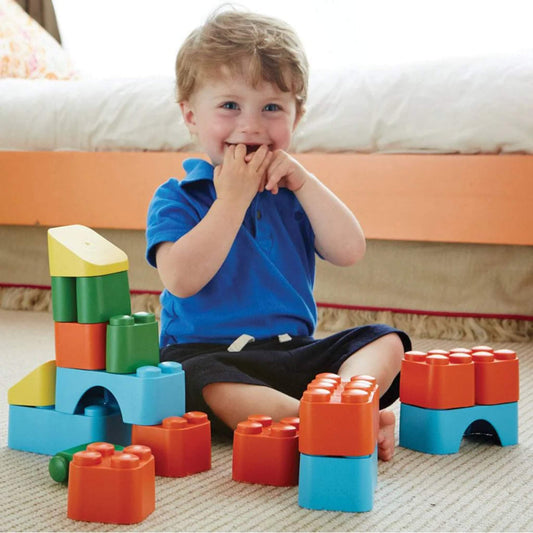 Young child sitting on the floor smiling beside a colourful block tower, surrounded by large interlocking blocks.