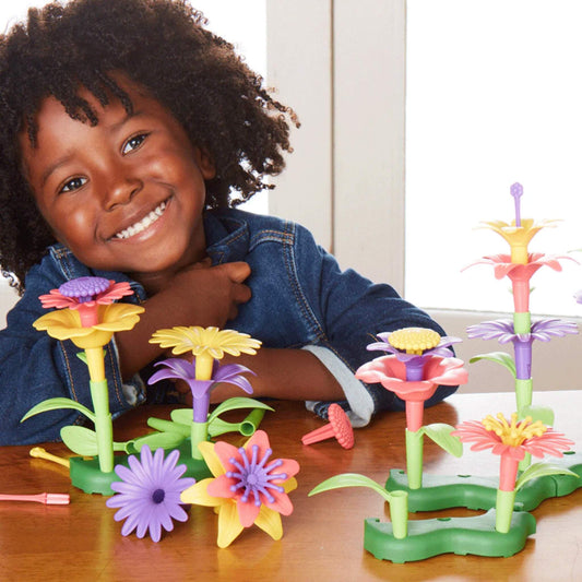 Child smiling at a table surrounded by colourful buildable flowers in pink, yellow, and purple with stems and leaves arranged in green bases.
