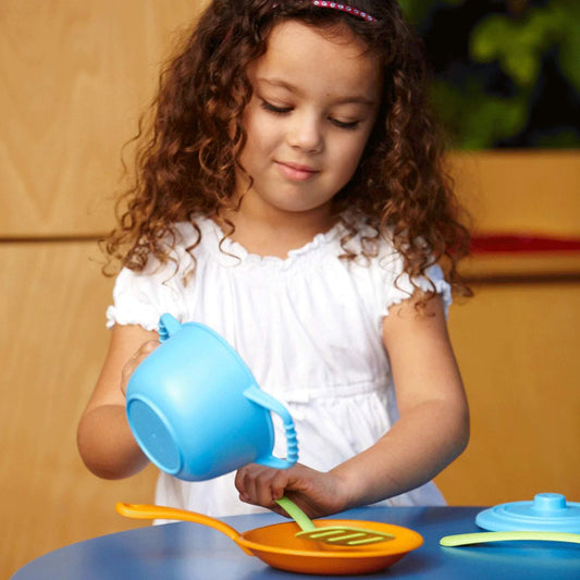 A young girl in a white dress stirring an orange frying pan with a green spatula while holding a blue pot, part of the Green Toys Chef Set.