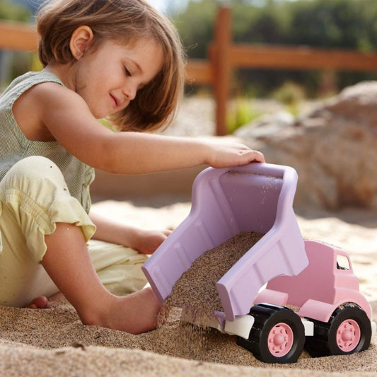 Young child sitting in a sandy outdoor play area, using the pink and purple dump truck to tip a load of sand over her foot while smiling and engaged.
