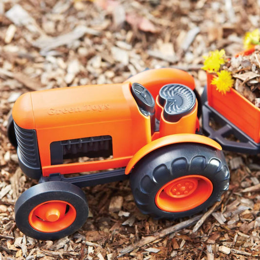 Toy tractor with rear trailer filled with bark and yellow flowers, placed outdoors on natural mulch surface.