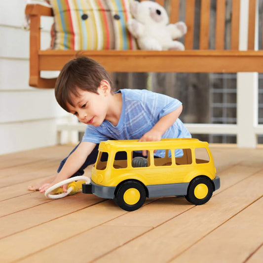 Young boy playing with the yellow school bus wagon on a wooden floor, holding the rope and reaching to guide it along.