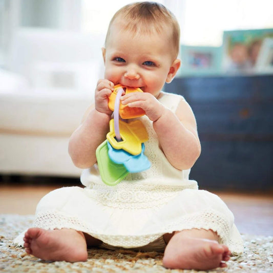 Smiling baby sitting on a rug, holding and mouthing the colourful Green Toys Rattle Keys, dressed in a white outfit in a softly lit room.