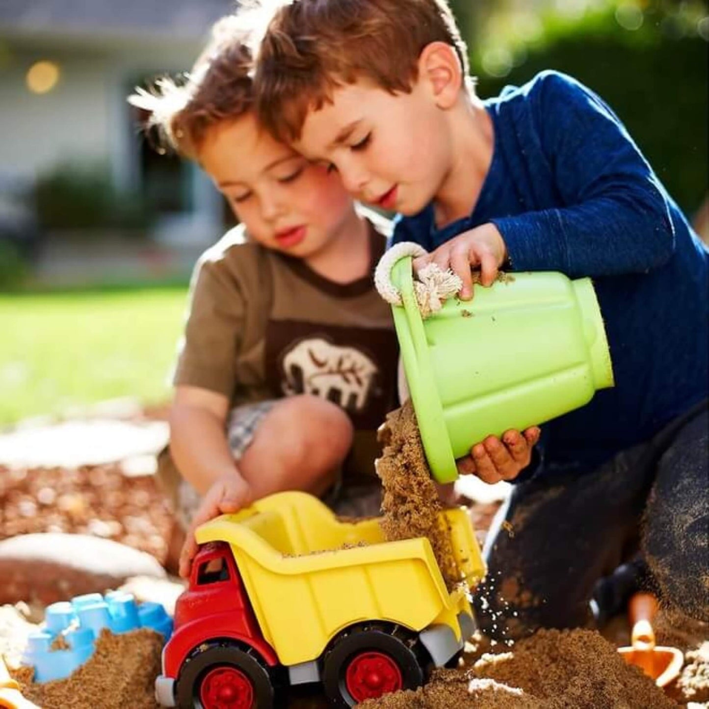 Two children outdoors filling a yellow toy dump truck with sand using the Green Toys green bucket, with a blue sand mould and orange tools visible in the sand.