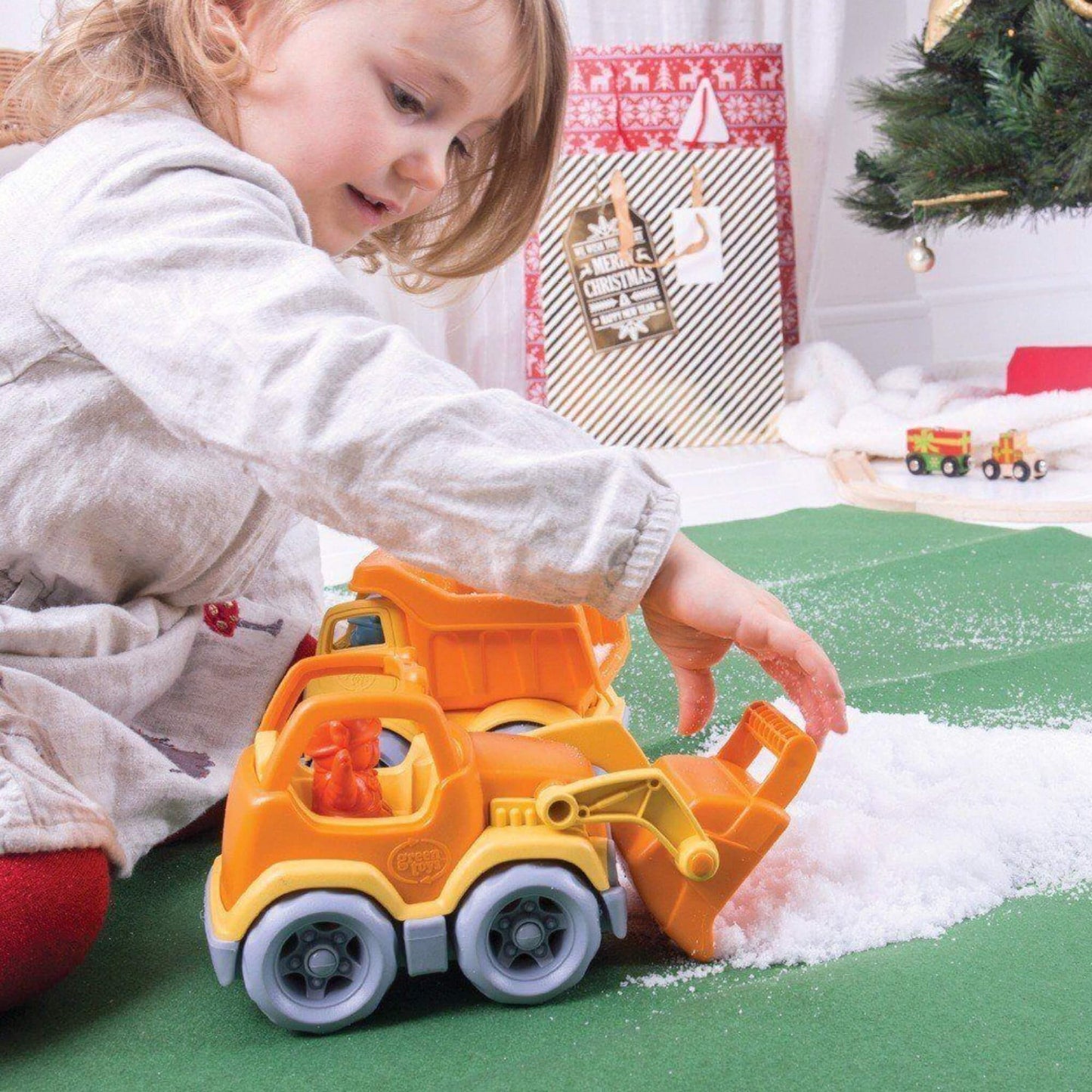 Toddler playing indoors on a green mat near a Christmas tree, using the scooper truck to shovel artificial snow in a festive play scene.