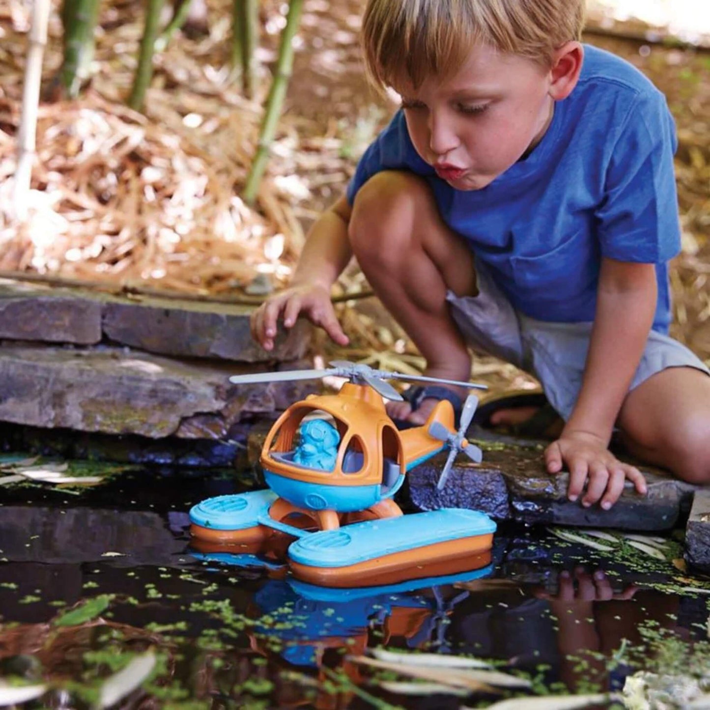 Child crouching by a garden pond, watching the Seacopter float on the surface among lily pads, with the pilot figure visible in the cockpit.
