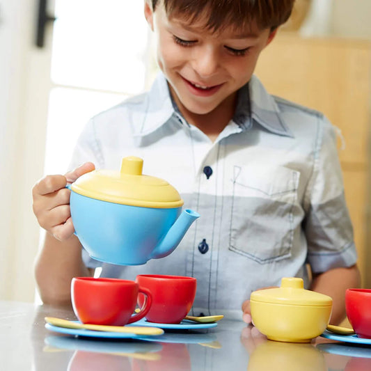 A smiling child pours from the blue and yellow Green Toys teapot into red cups on blue saucers, enjoying imaginative tea party play at a table.