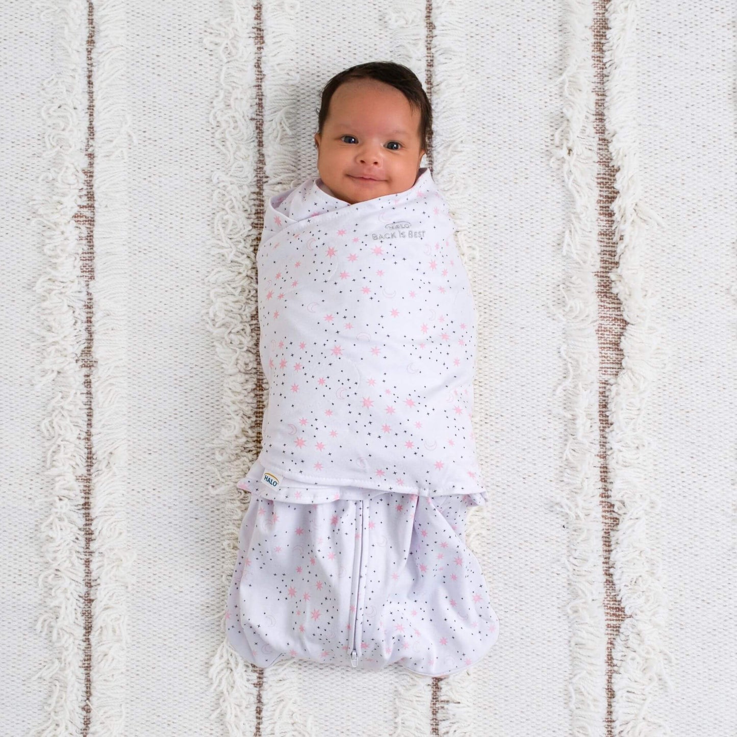 Smiling baby swaddled in HALO Midnight Moons Pink SleepSack, lying on a textured cream rug with arms tucked in for comfort.