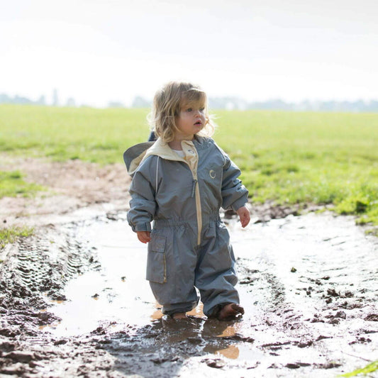 Toddler standing in a muddy puddle wearing a grey waterproof fleece lined suit with hood and beige zip, playing outdoors on a bright day.