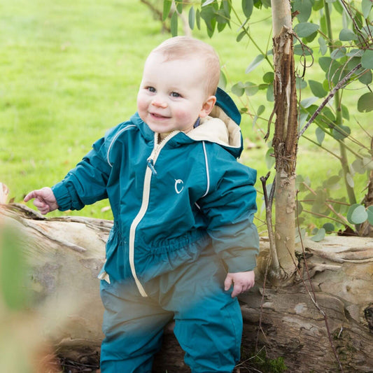 Smiling baby wearing a peacock green waterproof fleece-lined suit, standing next to a fallen tree outdoors in a grassy area.