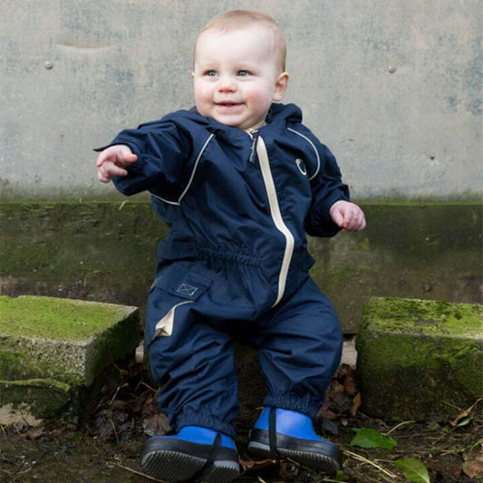 Baby sitting outdoors wearing a navy waterproof all-in-one suit with beige zip and hood, smiling and pointing while wearing blue wellies.