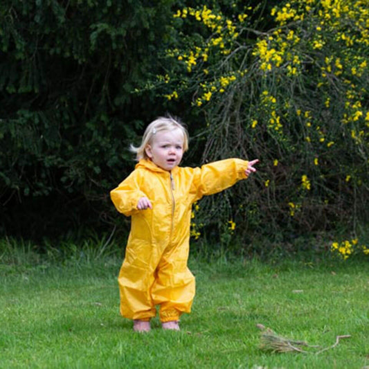 A toddler in a bright yellow rain suit stands on grass, pointing towards something with a curious expression, surrounded by greenery and yellow flowers.