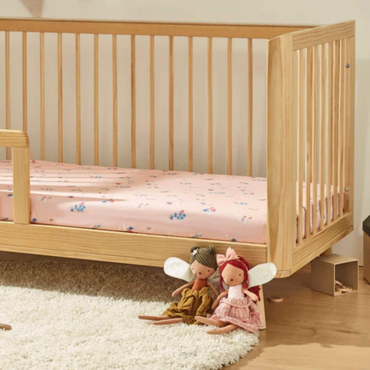 A wooden cot bed dressed with a pink floral fitted sheet, shown in a nursery setting with soft toys placed beside the cot.