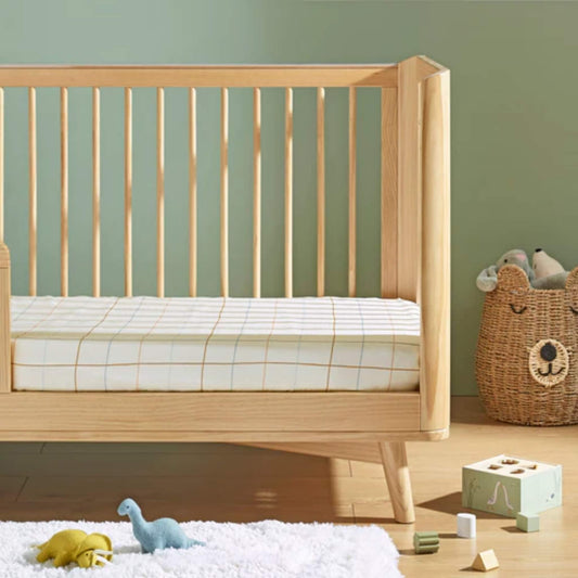 A wooden cot bed styled with a fitted sheet featuring a subtle grid pattern, placed in a calm nursery setting with soft toys and wooden blocks.