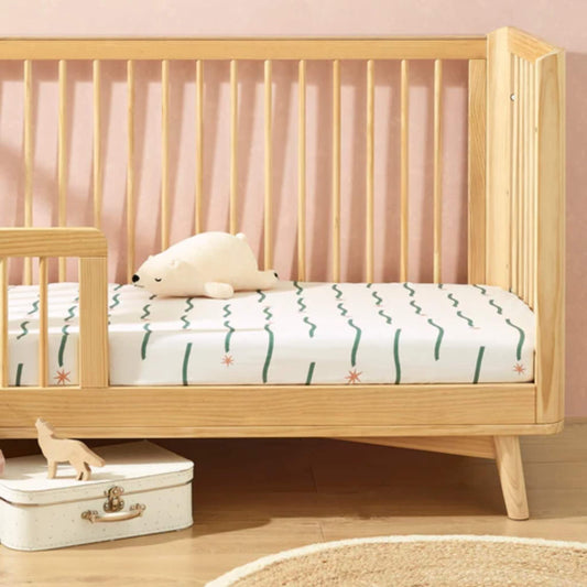 A cot bed dressed with a fitted sheet in a Night Sky print, featuring green wavy lines and star details against a warm wooden nursery setting.