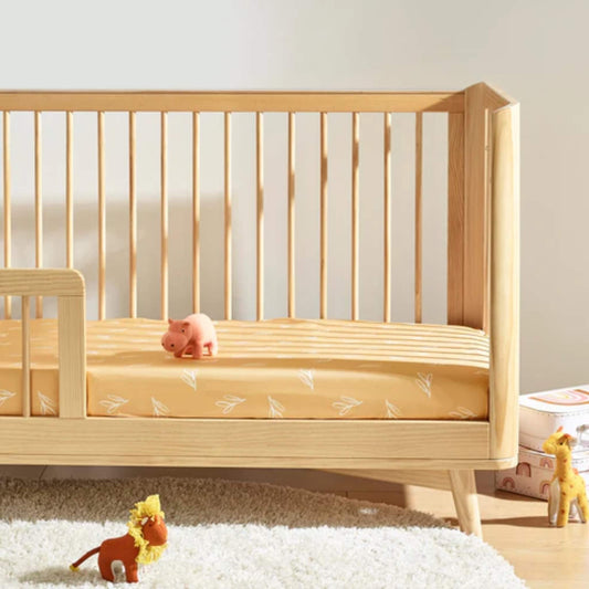 A wooden cot bed styled with a mustard fitted sheet featuring scattered white sprig illustrations, photographed in a softly lit nursery setting.