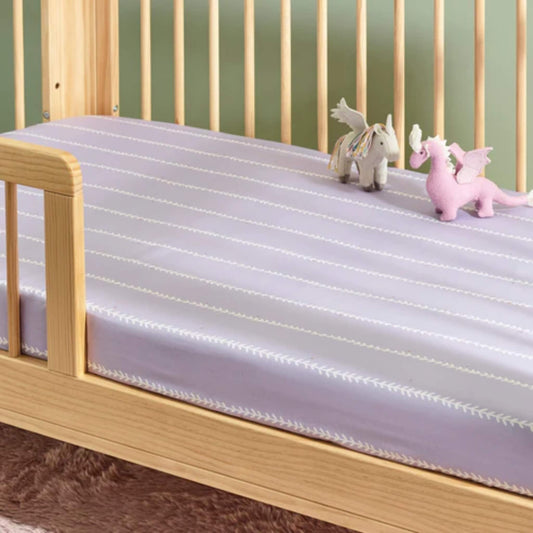 A lilac fitted sheet fitted on a wooden cot mattress with two small soft toys placed on top.