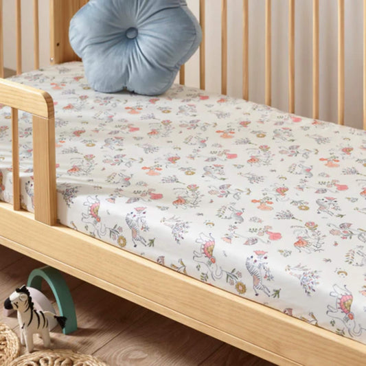 A fitted cotton sheet laid on a wooden cot bed, featuring a subtle boho-style print with animals, flowers, and soft muted colours.