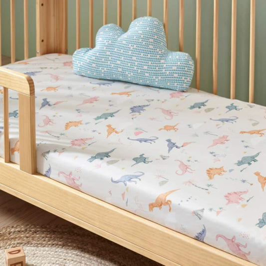 A fitted cotton sheet on a wooden cot bed, featuring a scattered dinosaur illustration in soft pastel colours across a light background.