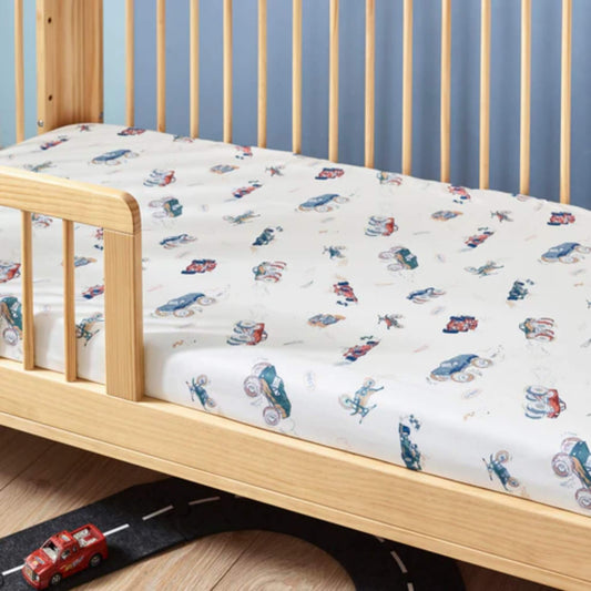 A fitted cotton sheet on a wooden cot bed, featuring a scattered vehicle pattern with illustrated cars, tractors, and bikes across a light background.
