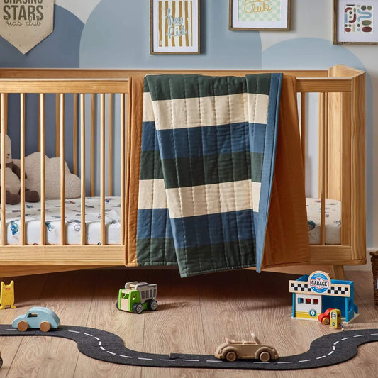 A wooden cot bed with a striped quilt draped over the side rail, showing wide colour-blocked stripes in blue, green, and cream with a warm ochre border.