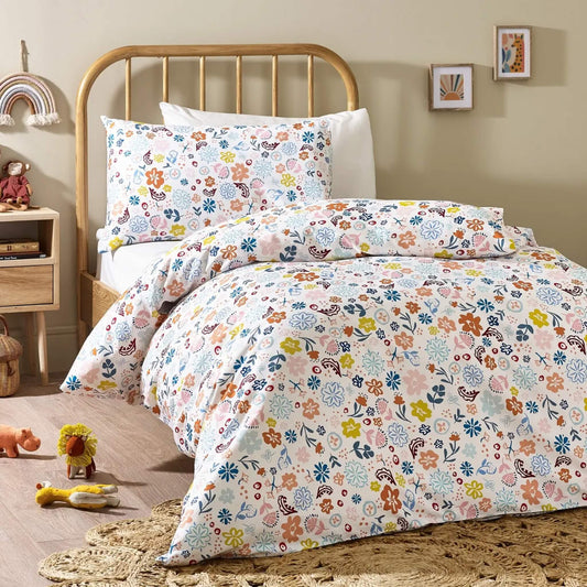 A full child's bed setup showing floral patterned bedding with layered pillows, combining pink, blue, yellow, and rust tones against a light cream background.