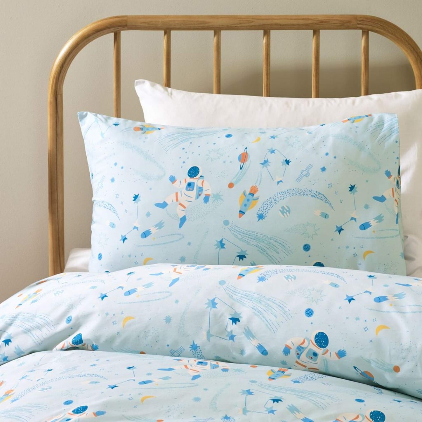 Close-up view of a pale blue pillowcase and duvet cover showing illustrated astronauts, rockets, stars and planets printed on soft cotton bedding.