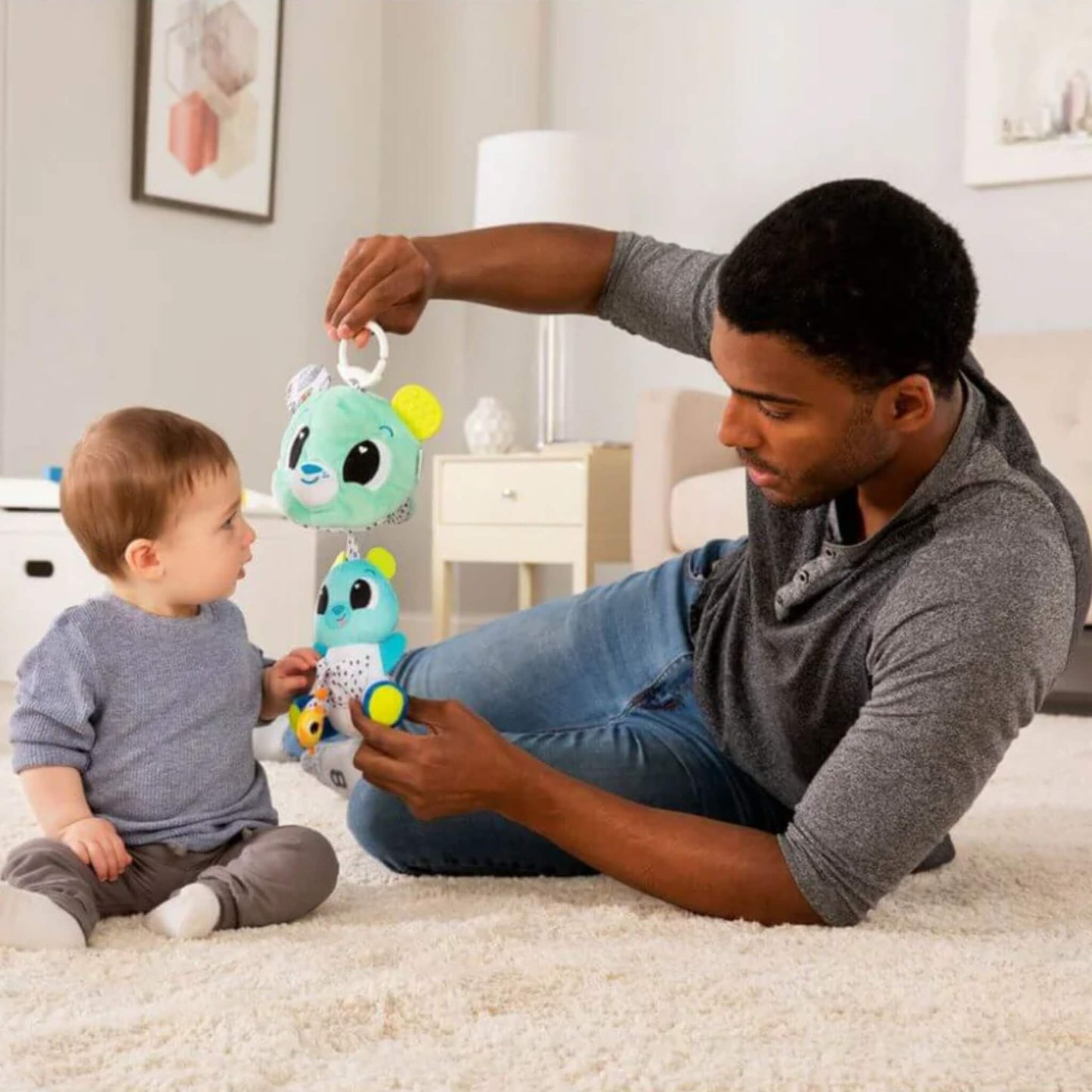 Father lying on a soft carpet interacting with his baby, holding a plush bear toy with layered bears for sensory discovery.