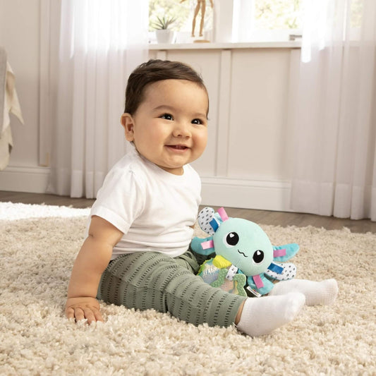 Smiling baby sitting on a soft cream carpet holding a plush axolotl toy with colourful textures and fins.