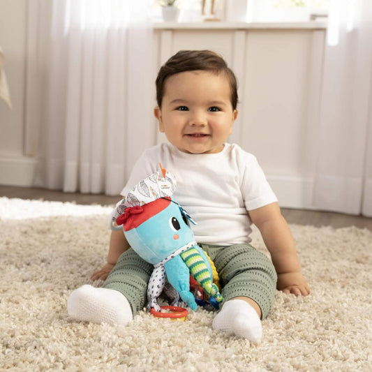 A baby sitting on a soft rug holding a colourful octopus-style activity toy with fabric legs and bright textures.