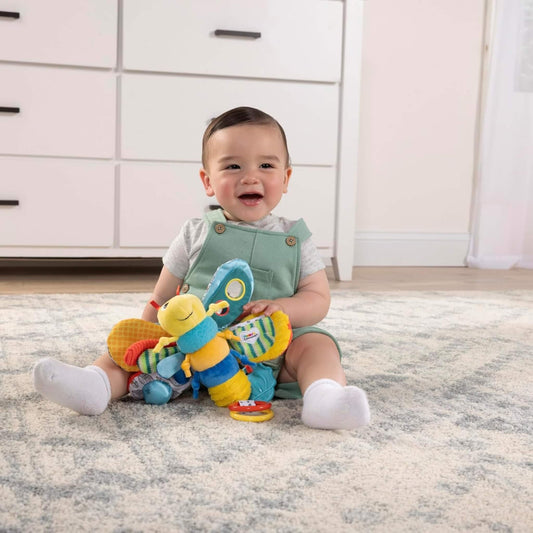 Baby sitting on a soft rug holding a plush firefly toy with large textured wings and bright patterns.