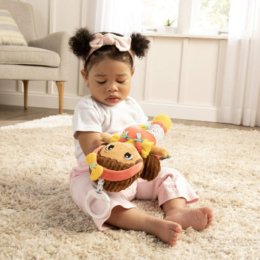 Baby sitting on a cream rug, holding a colourful soft doll with pigtails and bright clothing while exploring its textures and details.