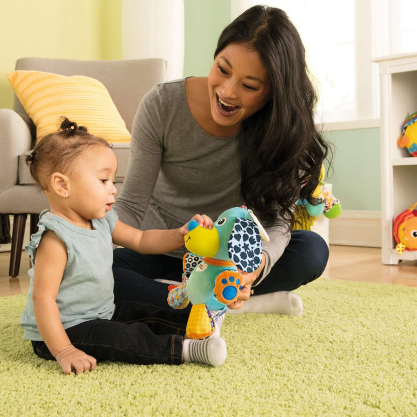Baby sitting on a green rug exploring the Pupsqueak toy with their mother during playtime.