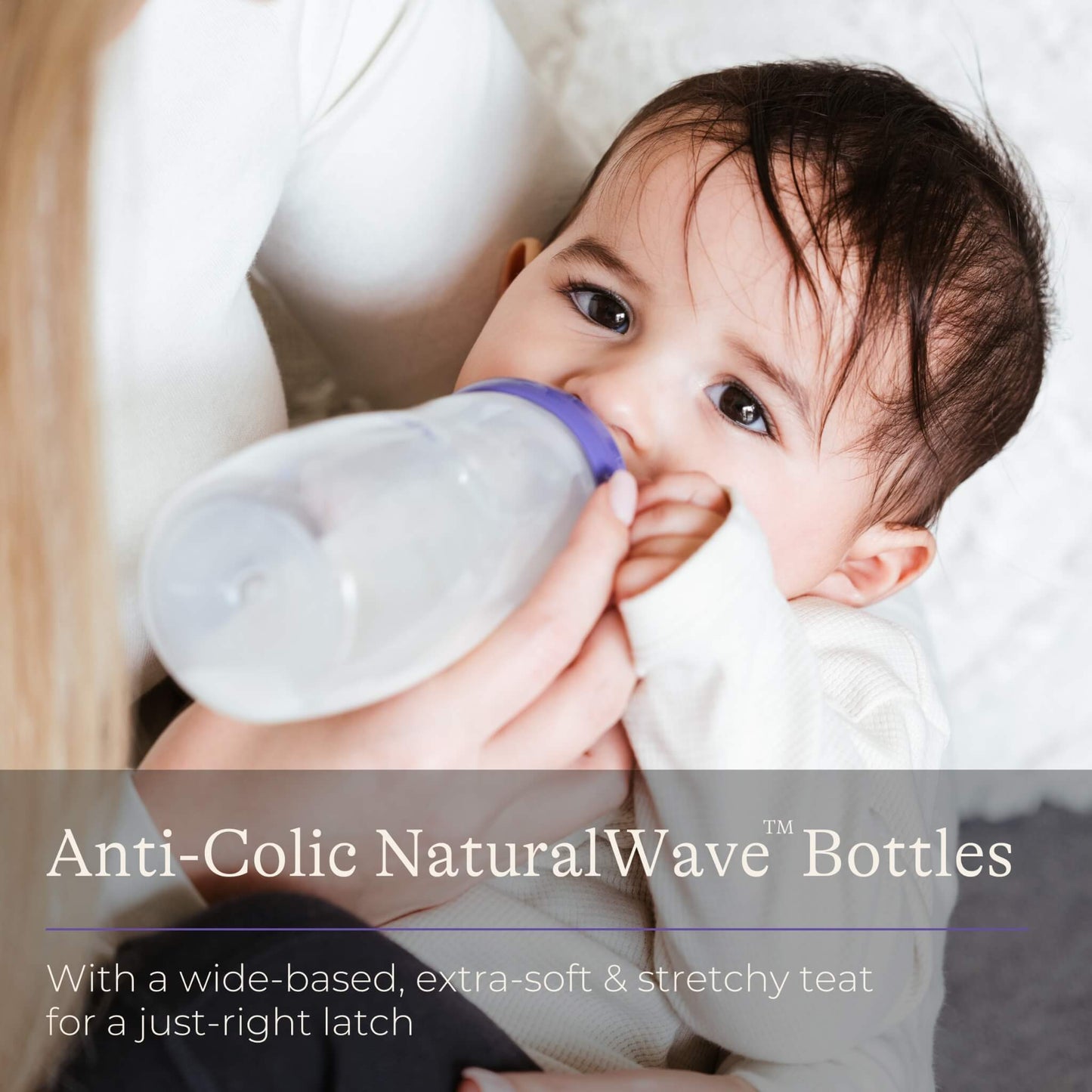 Baby being bottle fed while looking towards camera, drinking from a wide-neck feeding bottle while being held by an adult.
