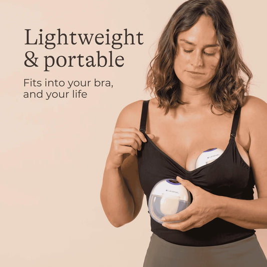 A woman positioning a Lansinoh wearable breast pump inside a black nursing bra while holding a second pump unit against her chest, demonstrating the compact wearable design.