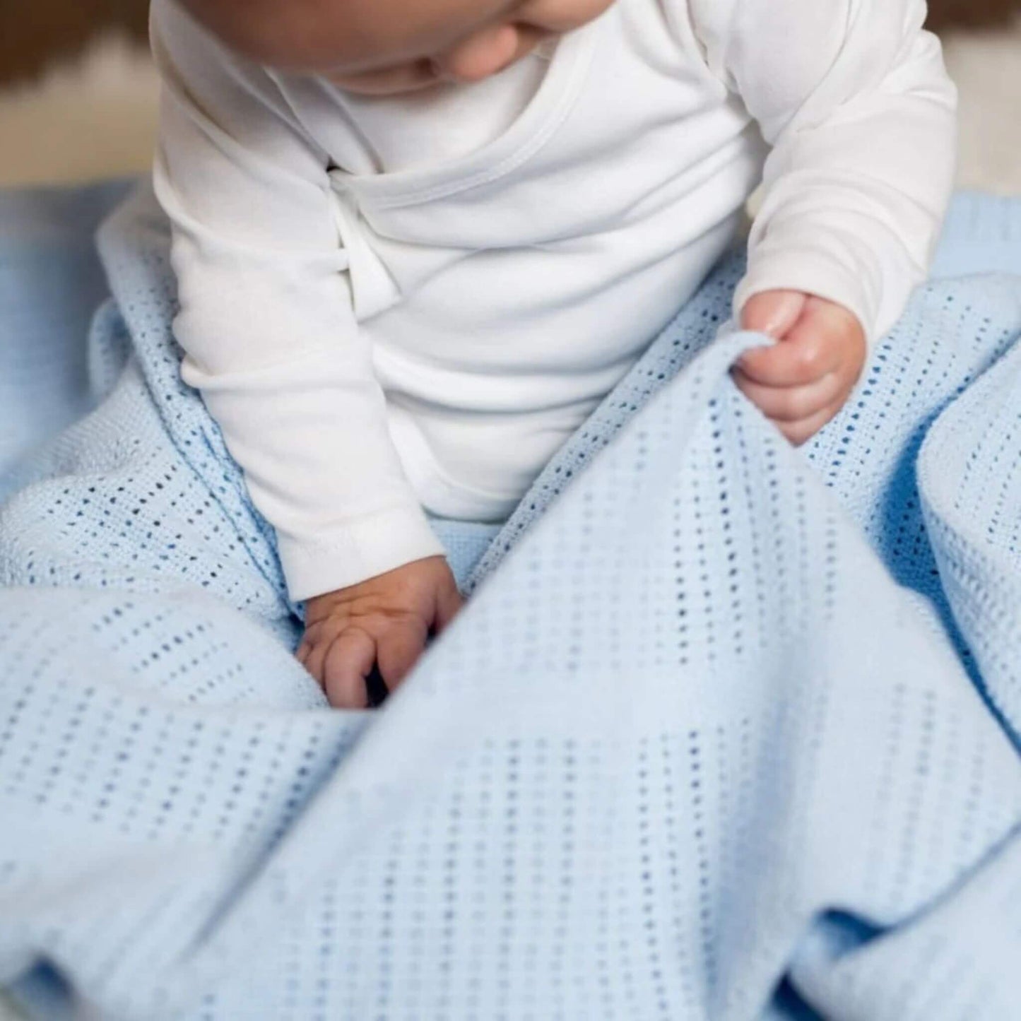Baby in white clothing snuggled with a light blue Lulujo cellular blanket, showing the cosy comfort.