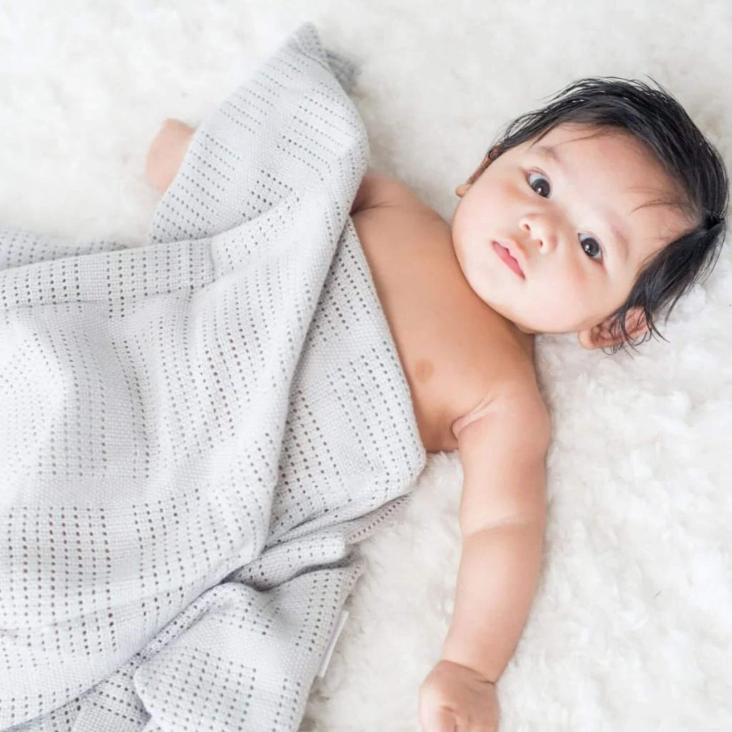 Peaceful baby resting on soft white bedding whilst partially covered with a light grey Lulujo cellular blanket, showcasing the breathable weave pattern and gentle comfort.
