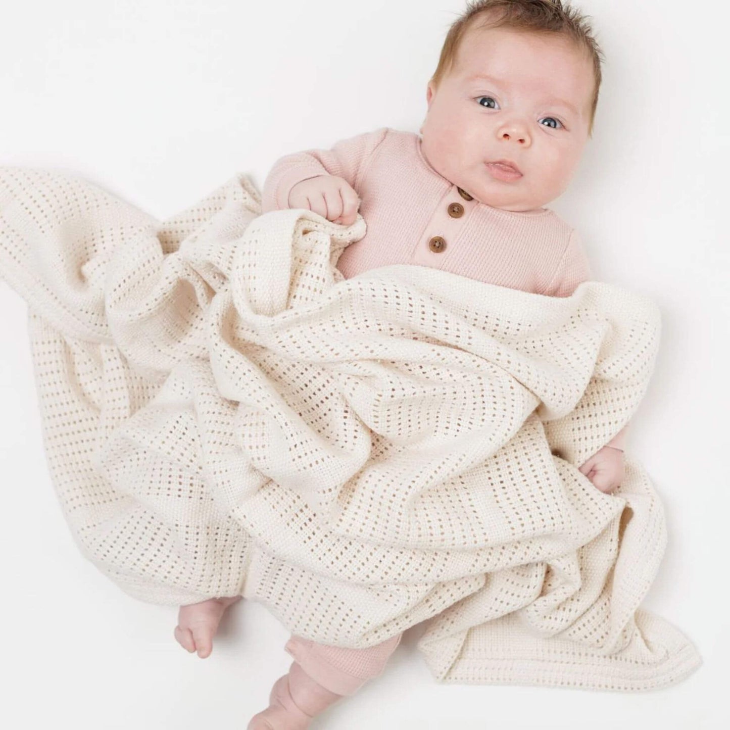 Baby in soft pink outfit loosely wrapped in cream-coloured Lulujo cellular blanket, showcasing the breathable weave pattern and versatile draping that allows perfect temperature regulation.