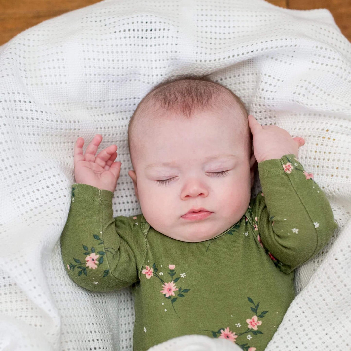 Peacefully sleeping baby in floral green outfit resting on a white Lulujo cellular blanket, highlighting the breathable fabric and soft texture perfect for promoting safe, comfortable sleep.