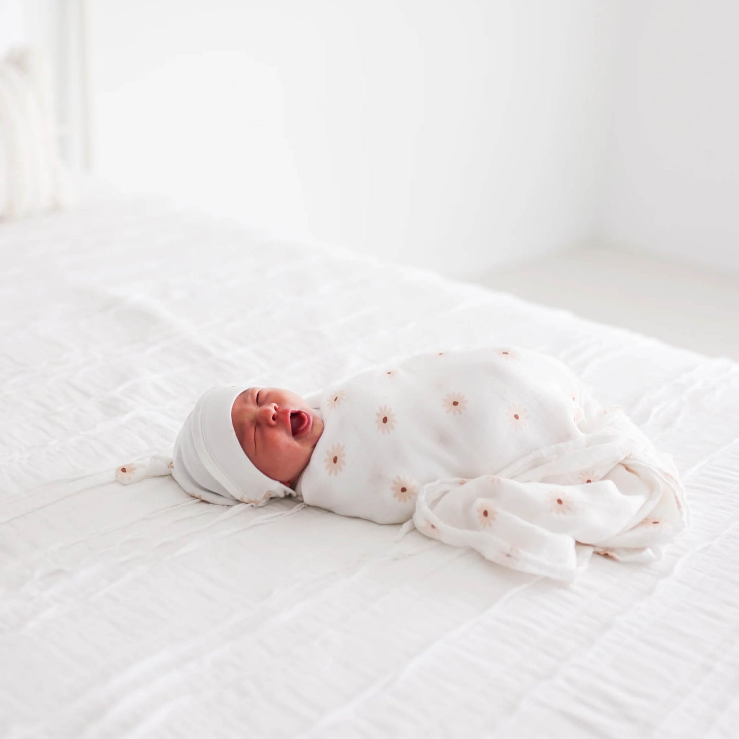 Newborn baby resting on a bed, wrapped in the white muslin swaddle decorated with soft peach daisies, captured mid-yawn.