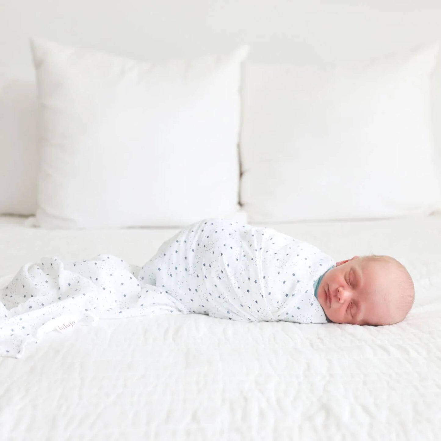 Baby sleeping on a white bed, wrapped in the starry muslin blanket with the Lulujo fabric tag visible at the edge.