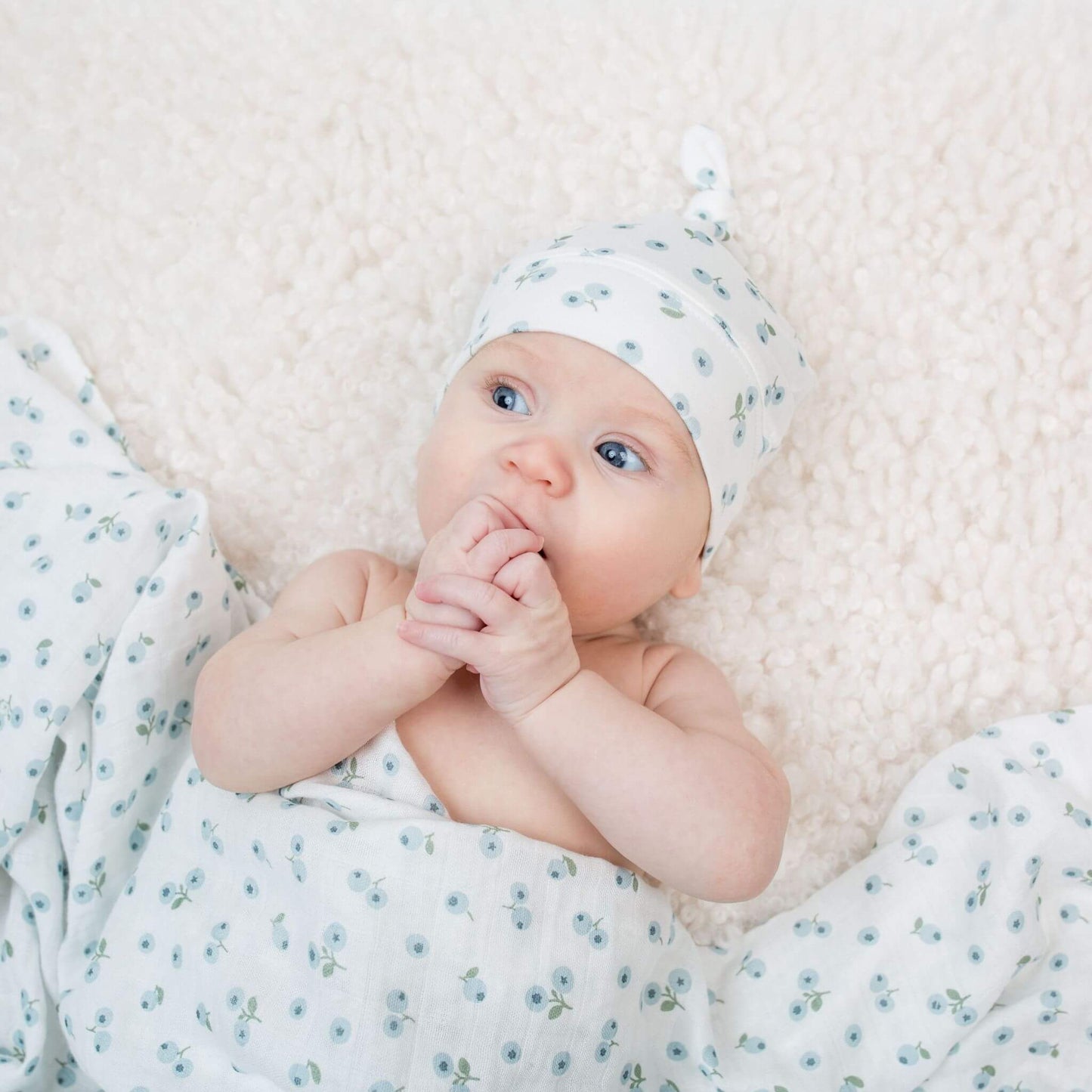Awake newborn baby with hand to mouth wearing Lulujo's blue floral knotted hat against white sherpa backdrop with matching muslin swaddle.
