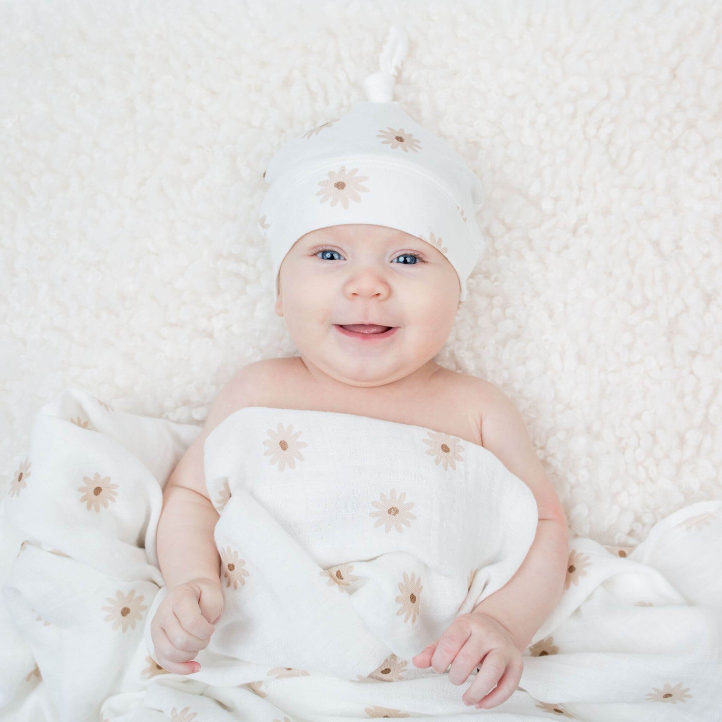 Smiling baby wearing Lulujo's white daisy print knotted hat with matching swaddle wrap, showcasing the soft fabric's comfort against delicate newborn skin.