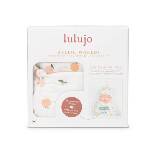 Lulujo Hello World set in packaging, featuring soft white muslin swaddle with peachy orange fruit pattern, matching knotted hat and personalized name tag for baby's first photos.