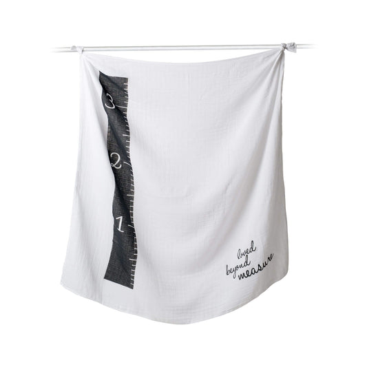 Lulujo Milestone Muslin Blanket displayed on hanging rod showing the black measuring tape design and "loved beyond measure" text for monthly baby photos.