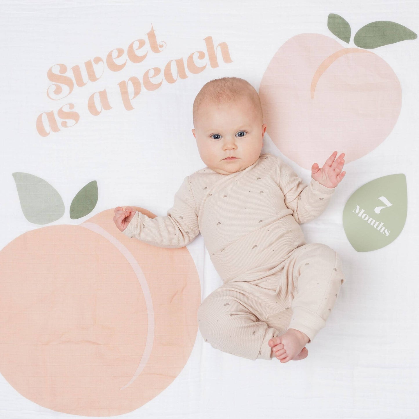 Seven-month-old baby in cream patterned outfit on Lulujo Sweet as a Peach milestone blanket with peach fruit designs and sage green milestone marker.