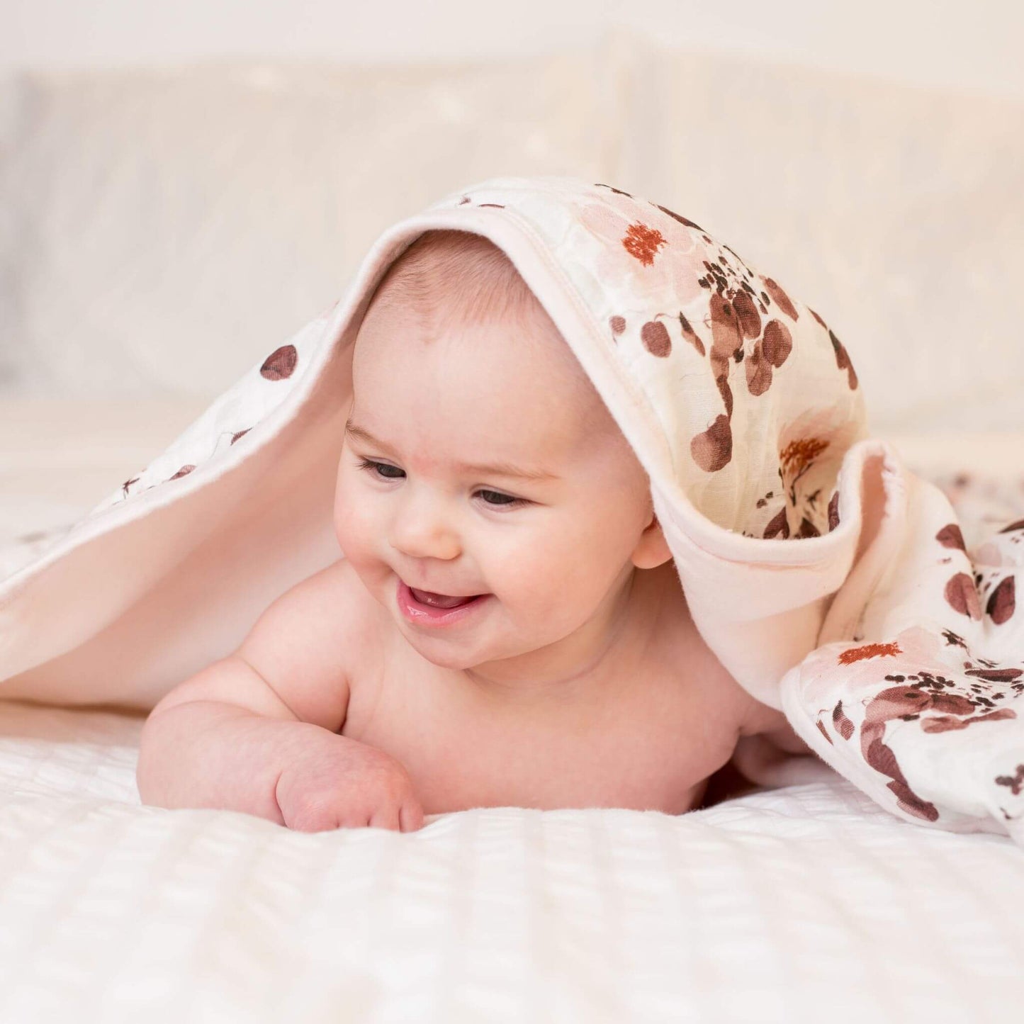 Smiling baby partially covered by the Lulujo eucalyptus print quilted blanket, showing how the soft fabric drapes comfortably while the baby enjoys tummy time.